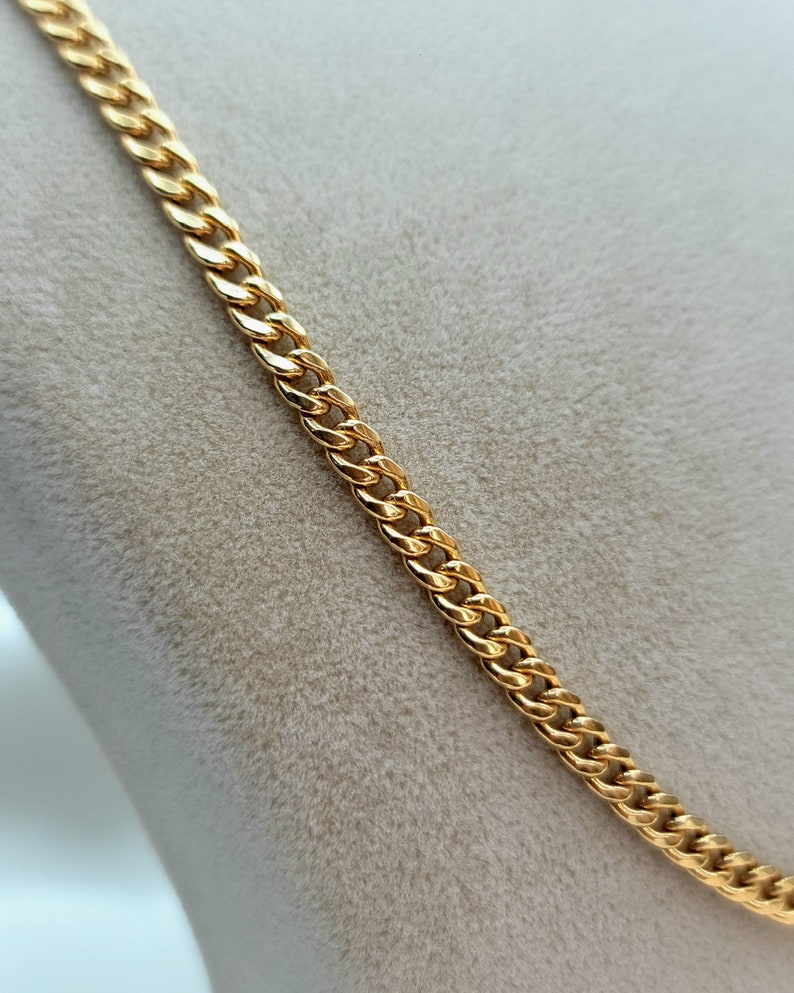 May include: A gold-colored curb chain necklace. The chain is made of interlocking links, creating a textured surface. The necklace is displayed against a neutral background, highlighting its shiny, metallic appearance. The chain is a classic jewelry piece.