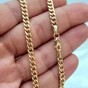 May include: A gold-colored curb chain necklace with a lobster clasp. The chain is made of interlocking links and is displayed against a light background. The clasp is marked with the number 750.