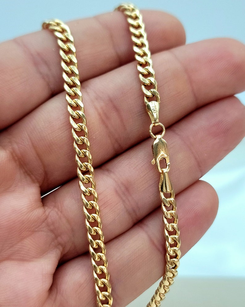 May include: A gold-colored curb chain necklace with a lobster clasp. The chain is made of interlocking links and is displayed against a light background. The clasp is marked with the number 750.