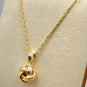 May include: A gold-colored necklace with a knot pendant. The pendant is a detailed knot design, and the chain is delicate. The necklace is displayed on a neutral-toned surface, highlighting the jewelry's shine and intricate details.