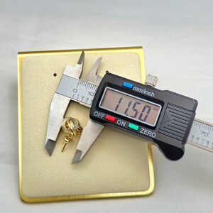 May include: A gold-colored knot-shaped earring is measured with a digital caliper, displaying 11.50 mm. The earring is on a beige display card with a gold border. The caliper is black with a digital display. The text "GoldLux Galleria" is in the bottom right corner.