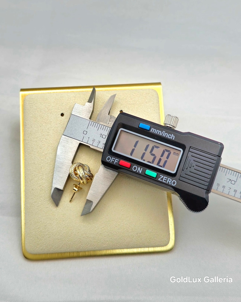 May include: A gold-colored knot-shaped earring is measured with a digital caliper, displaying 11.50 mm. The earring is on a beige display card with a gold border. The caliper is black with a digital display. The text "GoldLux Galleria" is in the bottom right corner.