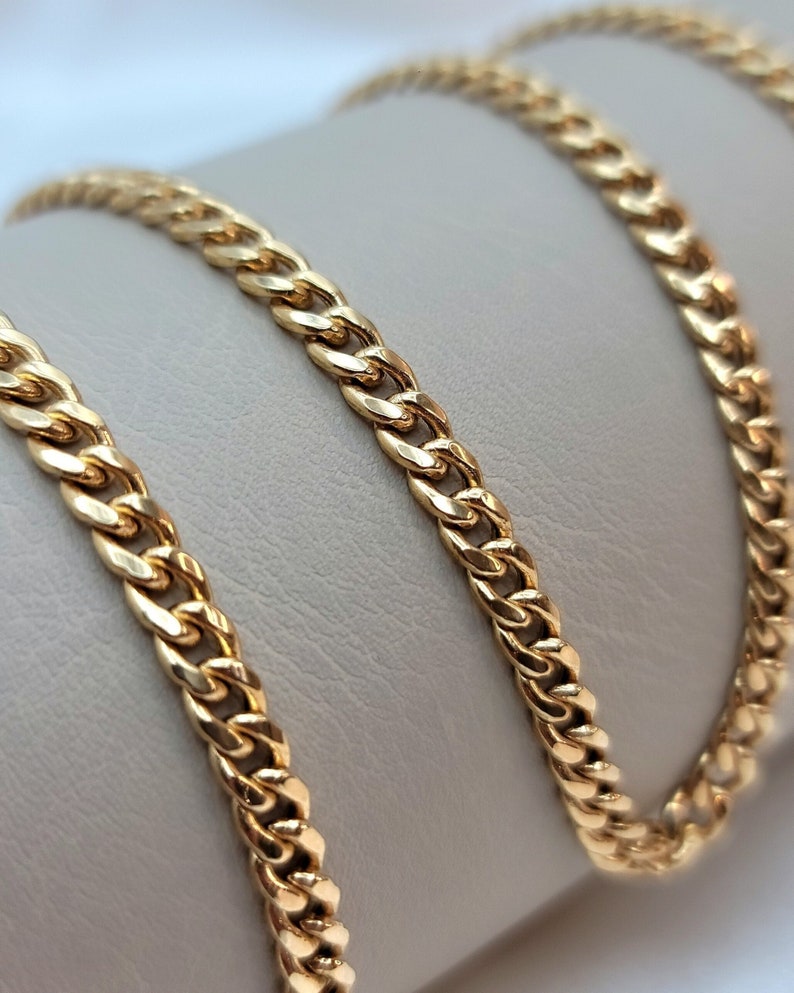 May include: A gold-colored curb chain necklace. The chain is made of interlocking links and is displayed on a neutral-toned surface. The chain has a polished finish, reflecting light.