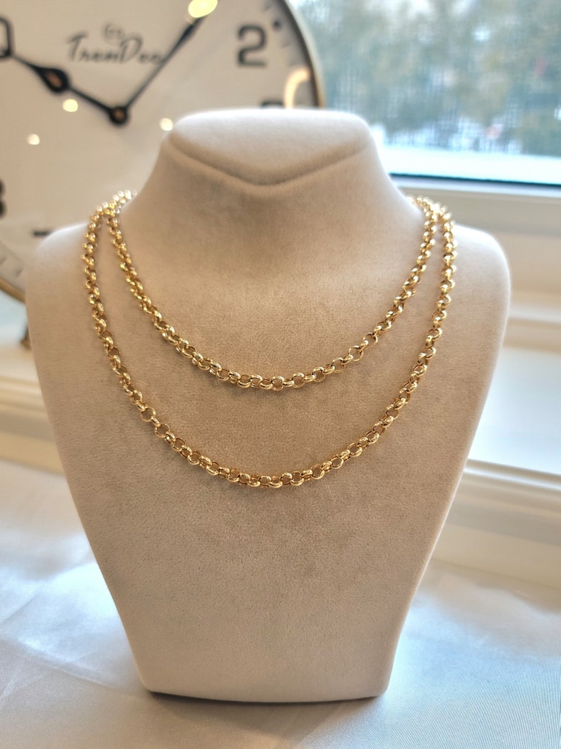 May include: Two gold chain necklaces layered on a white velvet display stand. The chains are made of small, round links.