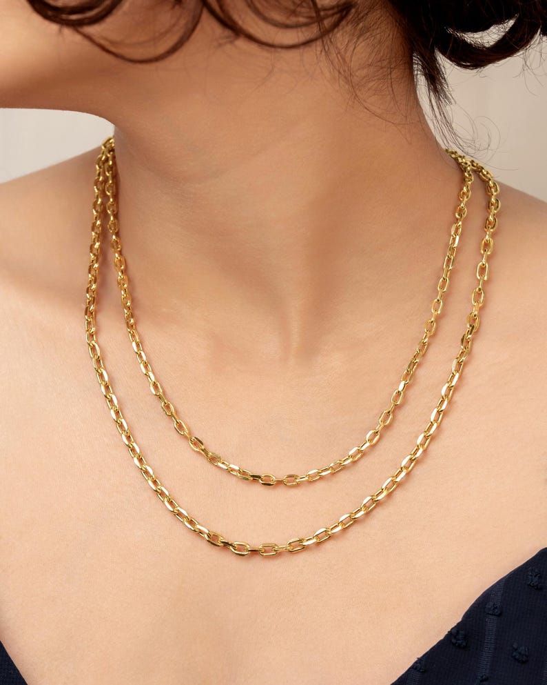 Puede incluir: Un collar de cadena de oro de doble capa con eslabones ovalados alargados. El collar consta de dos hilos de metal dorado pulido, con el hilo m&aacute;s corto descansando m&aacute;s arriba en el cuello y el m&aacute;s largo cayendo m&aacute;s abajo, creando un aspecto superpuesto.
