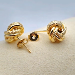 May include: A pair of gold-colored knot stud earrings. The earrings have a textured, woven design and are displayed with their backs and posts visible. The background is a light beige color, with a soft, blurred backdrop.