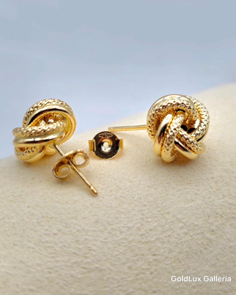 May include: A pair of gold-colored knot stud earrings. The earrings have a textured, woven design and are displayed with their backs and posts visible. The background is a light beige color, with a soft, blurred backdrop.