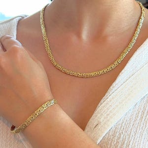 May include: A gold-coloured necklace and bracelet set. The jewellery features a detailed, textured design. The necklace is around the neck, and the bracelet is on the wrist. The jewellery is worn over a white top.