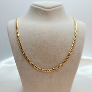 May include: A gold-colored curb chain necklace displayed on a beige jewelry bust. The chain has a classic, linked design and a bright, polished finish. The necklace is a simple yet elegant piece of jewelry.