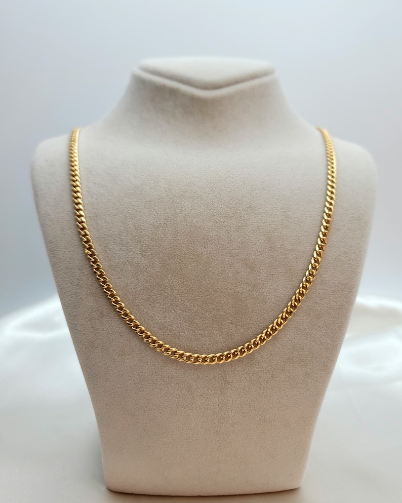 May include: A gold-colored curb chain necklace displayed on a beige jewelry bust. The chain has a classic, linked design and a bright, polished finish. The necklace is a simple yet elegant piece of jewelry.