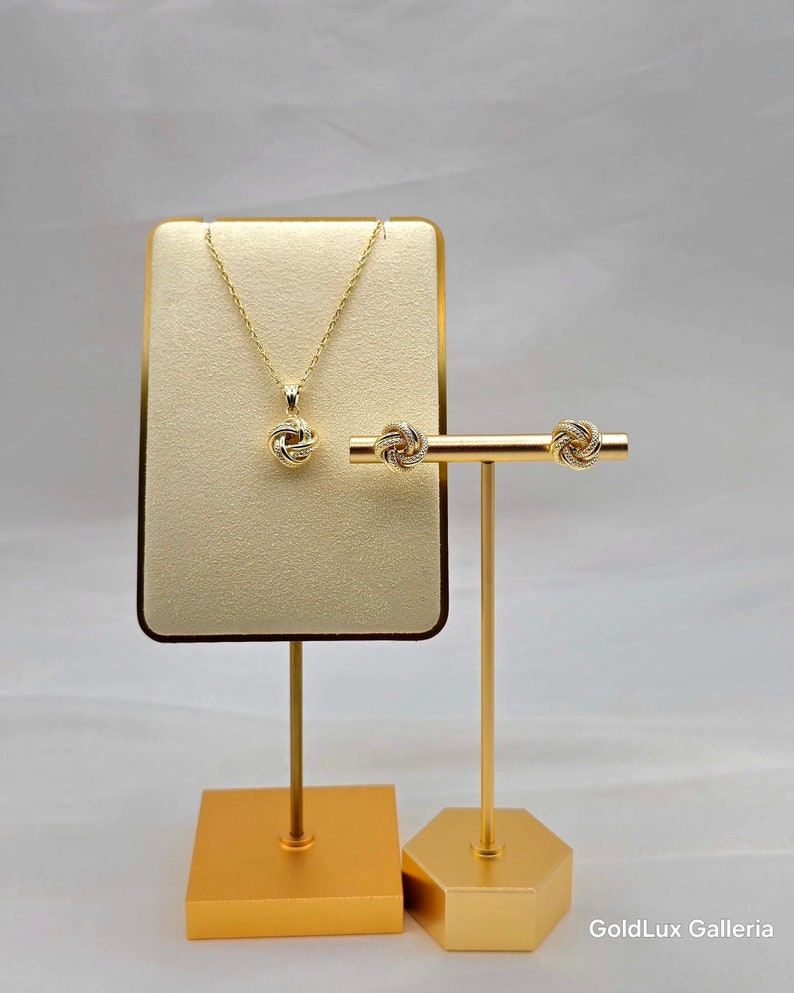 May include: A gold-colored necklace and matching earrings set displayed on a gold stand. The necklace features a knot pendant, and the earrings have a similar knot design. The jewelry is presented on a beige backdrop, with the brand name "GoldLux Galleria" visible.