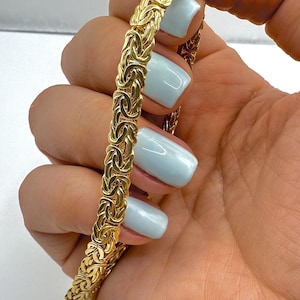 May include: A gold-coloured Byzantine chain bracelet. The bracelet is held up against a light background. The bracelet has a detailed woven design and a secure clasp. The bracelet is a jewelry item.