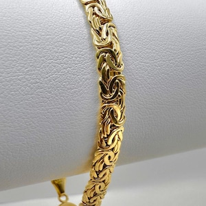 May include: A gold-coloured Byzantine bracelet. The bracelet has a detailed, woven design with a secure clasp. The jewellery is displayed against a white background, highlighting its intricate pattern and warm hue.