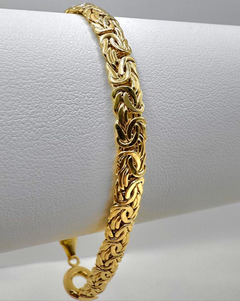 May include: A gold-coloured Byzantine bracelet. The bracelet has a detailed, woven design with a secure clasp. The jewellery is displayed against a white background, highlighting its intricate pattern and warm hue.