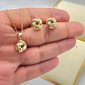 May include: A gold-colored jewelry set featuring a pendant necklace and matching stud earrings. The pendant and earrings have a knot design with small, sparkling accents. The necklace chain is delicate, and the set is displayed on a hand.
