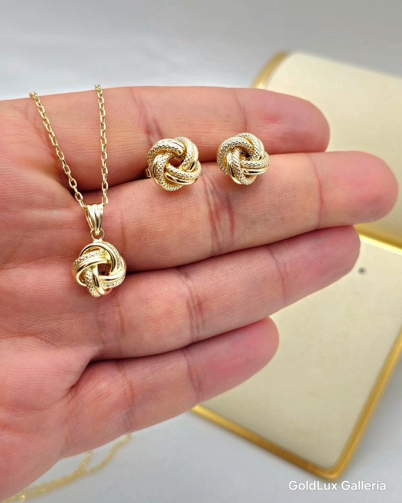 May include: A gold-colored jewelry set featuring a pendant necklace and matching stud earrings. The pendant and earrings have a knot design with small, sparkling accents. The necklace chain is delicate, and the set is displayed on a hand.