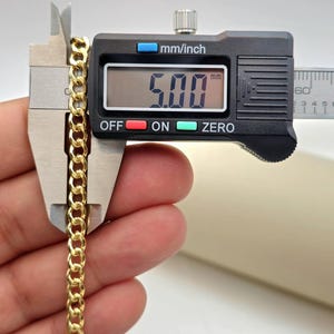 May include: A gold-colored curb chain is measured with a digital caliper. The caliper displays a measurement of 5.00 mm. The chain is held between the caliper jaws, showcasing its linked design. The background is a neutral color.