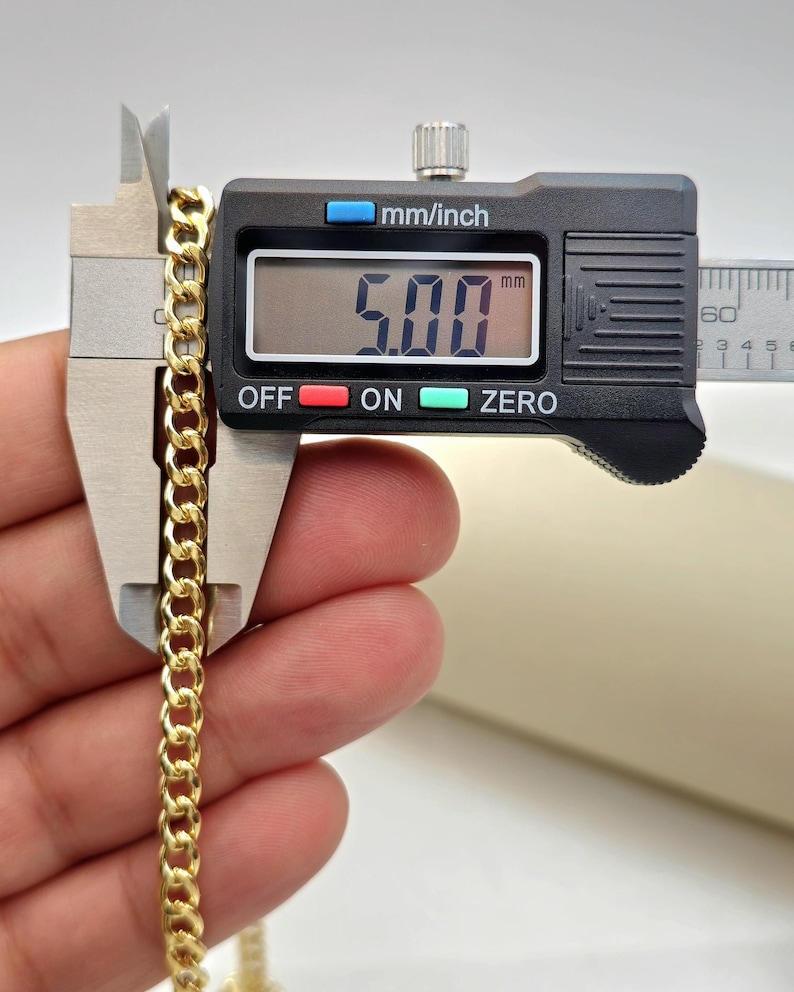 May include: A gold-colored curb chain is measured with a digital caliper. The caliper displays a measurement of 5.00 mm. The chain is held between the caliper jaws, showcasing its linked design. The background is a neutral color.