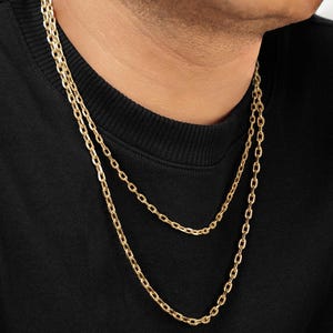 Puede incluir: Un collar de cadena doble en oro con eslabones ovalados alargados. El collar se lleva alrededor del cuello, sobre una prenda negra. El acabado dorado met&aacute;lico refleja la luz, resaltando la forma uniforme de cada eslab&oacute;n individual.