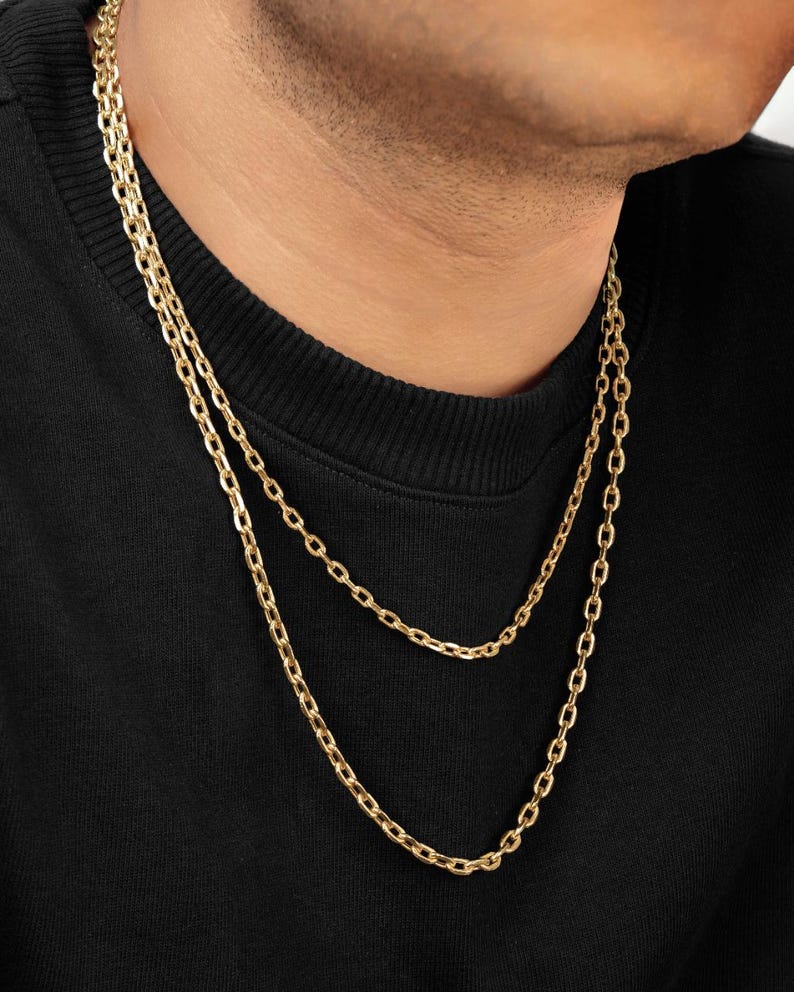 Puede incluir: Un collar de cadena doble en oro con eslabones ovalados alargados. El collar se lleva alrededor del cuello, sobre una prenda negra. El acabado dorado met&aacute;lico refleja la luz, resaltando la forma uniforme de cada eslab&oacute;n individual.