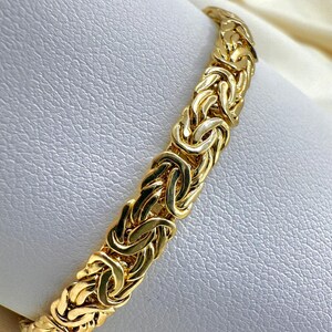 May include: A gold Byzantine chain bracelet. The bracelet has a woven design with interlocking links. The jewellery is displayed on a white surface, highlighting the intricate details and the warm tones of the metal.