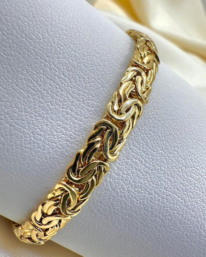 May include: A gold Byzantine chain bracelet. The bracelet has a woven design with interlocking links. The jewellery is displayed on a white surface, highlighting the intricate details and the warm tones of the metal.