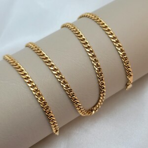May include: A gold-colored curb chain necklace displayed on a neutral-toned surface. The chain is made of interlocking links, creating a textured appearance. The necklace is shown in a curved arrangement, highlighting its design and shine.
