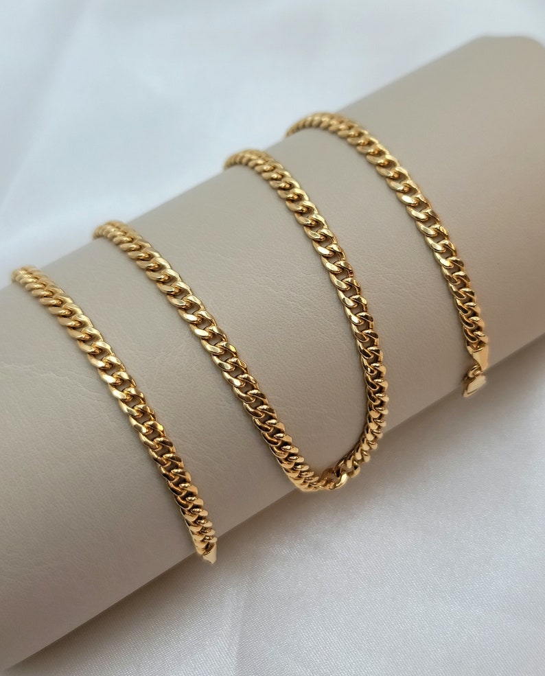 May include: A gold-colored curb chain necklace displayed on a neutral-toned surface. The chain is made of interlocking links, creating a textured appearance. The necklace is shown in a curved arrangement, highlighting its design and shine.