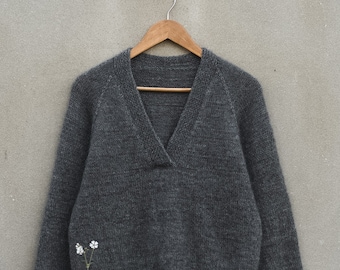 May include: A gray knitted sweater with a v-neck and long sleeves hangs on a wooden hanger against a gray wall.