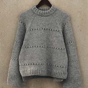 Könnte beinhalten: Ein grauer Strickpullover mit Rundhalsausschnitt und langen Ärmeln. Der Pullover hat ein zartes Lochmuster.