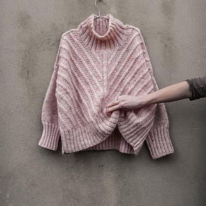 May include: A light pink, oversized sweater with a high, ribbed collar and ribbed cuffs. The sweater is hanging on a silver hanger against a gray wall. The sweater has a textured, diagonal knit pattern.