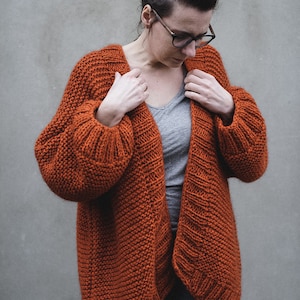 May include: A rust-colored, chunky knit cardigan with a relaxed fit. The open-front sweater features ribbed cuffs and a textured stitch pattern. The model is wearing a gray t-shirt and blue jeans. The cardigan is a cozy and stylish garment.