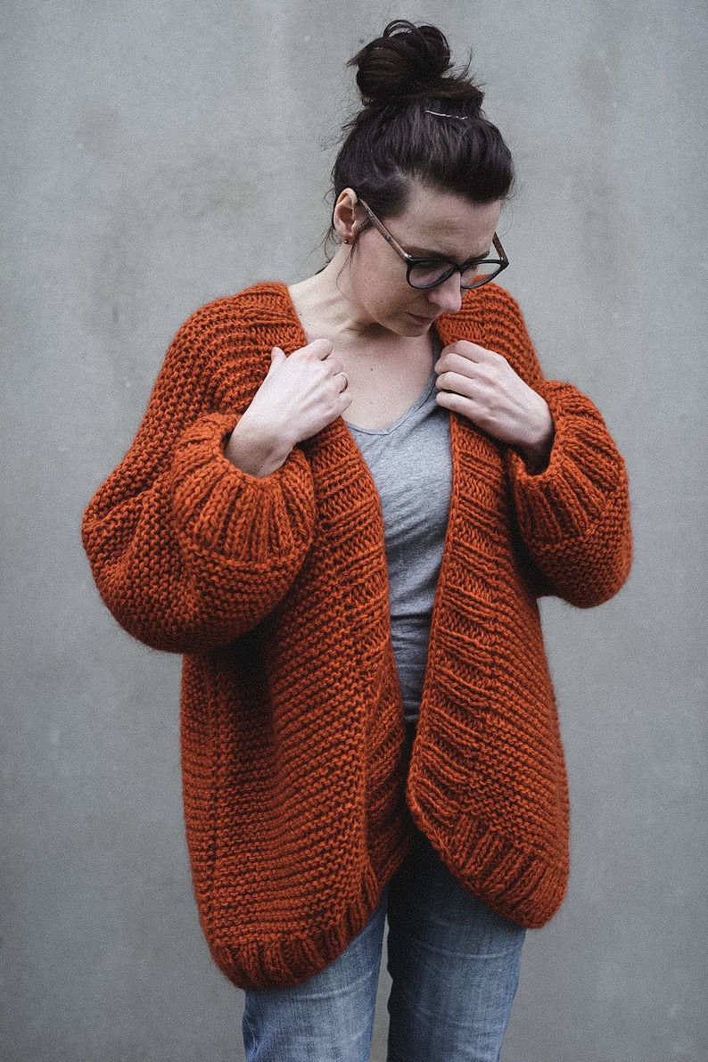 May include: A rust-colored, chunky knit cardigan with a relaxed fit. The open-front sweater features ribbed cuffs and a textured stitch pattern. The model is wearing a gray t-shirt and blue jeans. The cardigan is a cozy and stylish garment.
