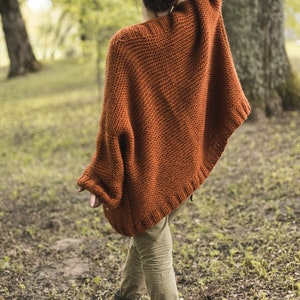 May include: A rust-colored, knitted poncho with wide sleeves. The poncho is worn over olive-green pants and black boots. The person is standing in a wooded area with trees and foliage in the background.
