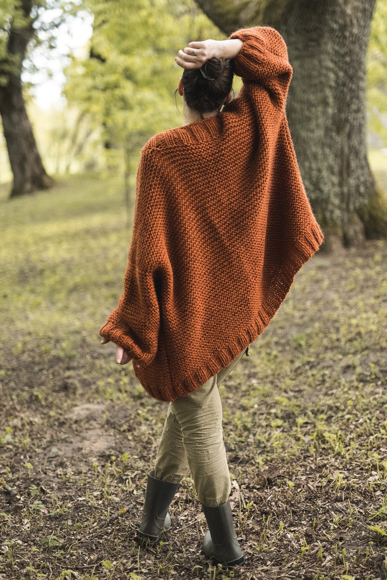 May include: A rust-colored, knitted poncho with wide sleeves. The poncho is worn over olive-green pants and black boots. The person is standing in a wooded area with trees and foliage in the background.