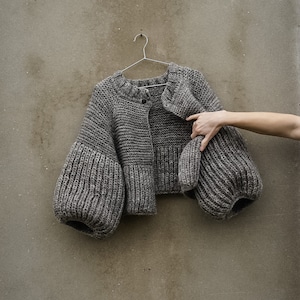 May include: A gray knitted cardigan with large, puffed sleeves. The cardigan has a button closure and a relaxed fit.