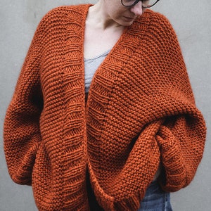May include: An oversized, rust-colored knit cardigan with a wide, open front. The chunky knitwear features a textured pattern and long sleeves. The cardigan is worn over a gray top and blue jeans, creating a cozy, casual look.