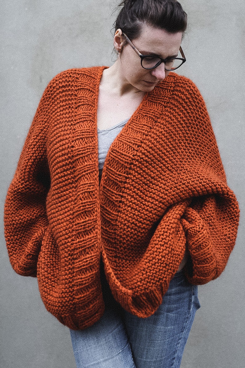 May include: An oversized, rust-colored knit cardigan with a wide, open front. The chunky knitwear features a textured pattern and long sleeves. The cardigan is worn over a gray top and blue jeans, creating a cozy, casual look.