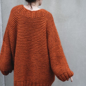 May include: An oversized, rust-colored knit sweater with a wide neckline and ribbed cuffs. The sweater has a chunky knit texture and long sleeves. The person is wearing blue jeans. The sweater is a cozy, warm garment.