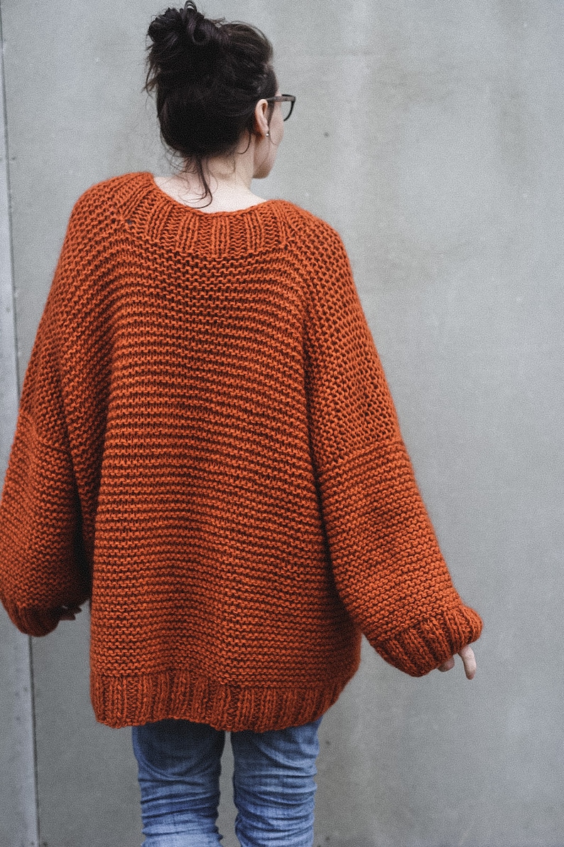 May include: An oversized, rust-colored knit sweater with a wide neckline and ribbed cuffs. The sweater has a chunky knit texture and long sleeves. The person is wearing blue jeans. The sweater is a cozy, warm garment.