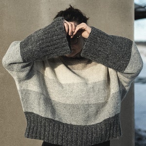 May include: A person wearing a gray, white, and black striped sweater with long sleeves. The sweater is made of a soft, knitted fabric.