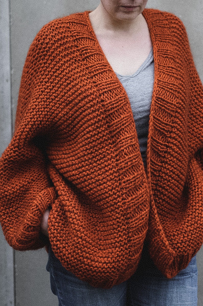 May include: A rust-colored, open-front cardigan with a chunky knit design. The sweater has a wide, relaxed fit with long sleeves. The garment is made of a thick, textured yarn, and is worn over a gray top and blue jeans.