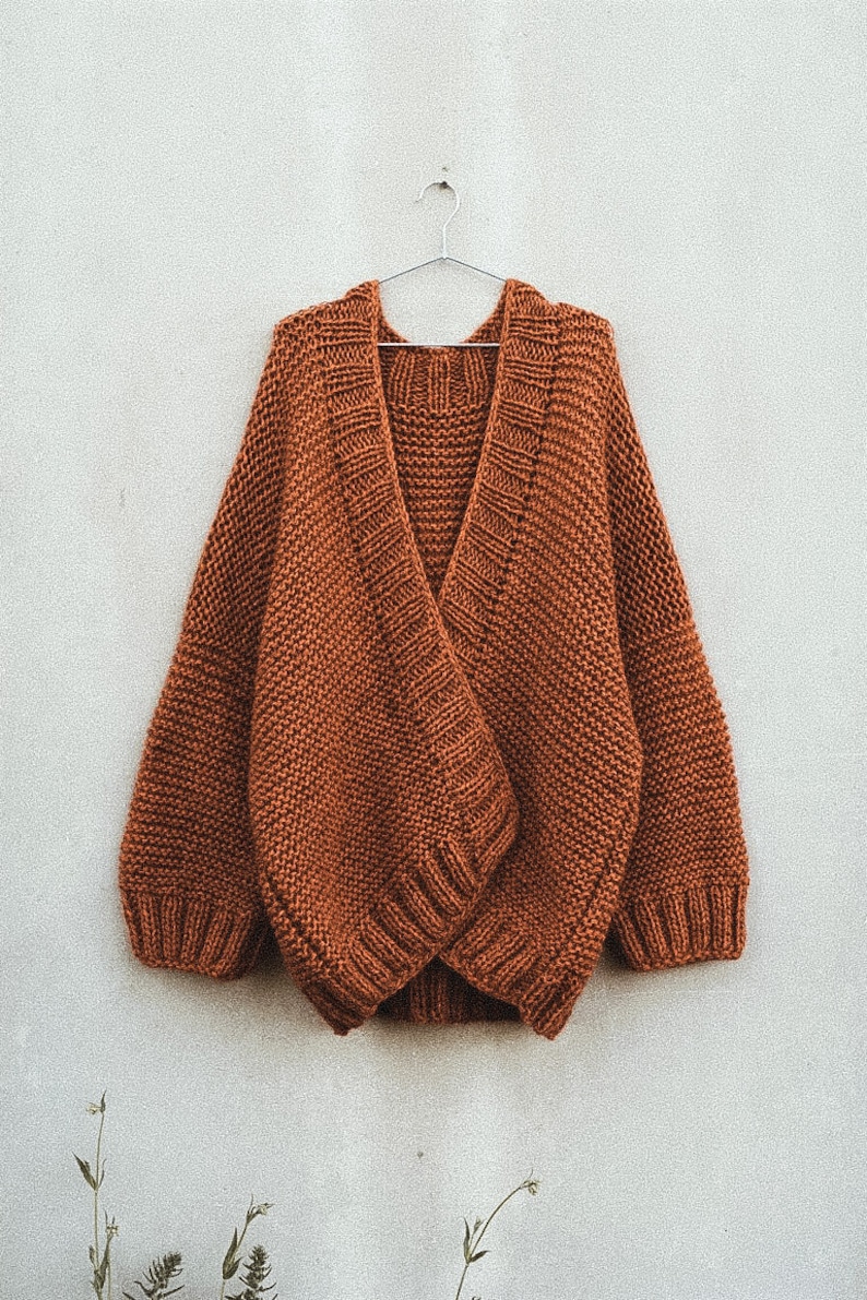 May include: A rust-colored, chunky knit cardigan hangs on a silver hanger against a white wall. The sweater features a deep V-neck and wide sleeves. The knit has a textured appearance.
