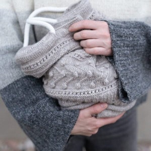 May include: A knitted bag with a white handle, featuring a textured pattern with small, raised circles. The bag is held by a person wearing a gray and white sweater.