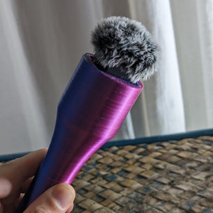 May include: A handheld microphone with a fluffy, gray windscreen. The microphone body transitions from blue to purple. The device is designed for recording audio. The background is a white curtain and a woven surface.