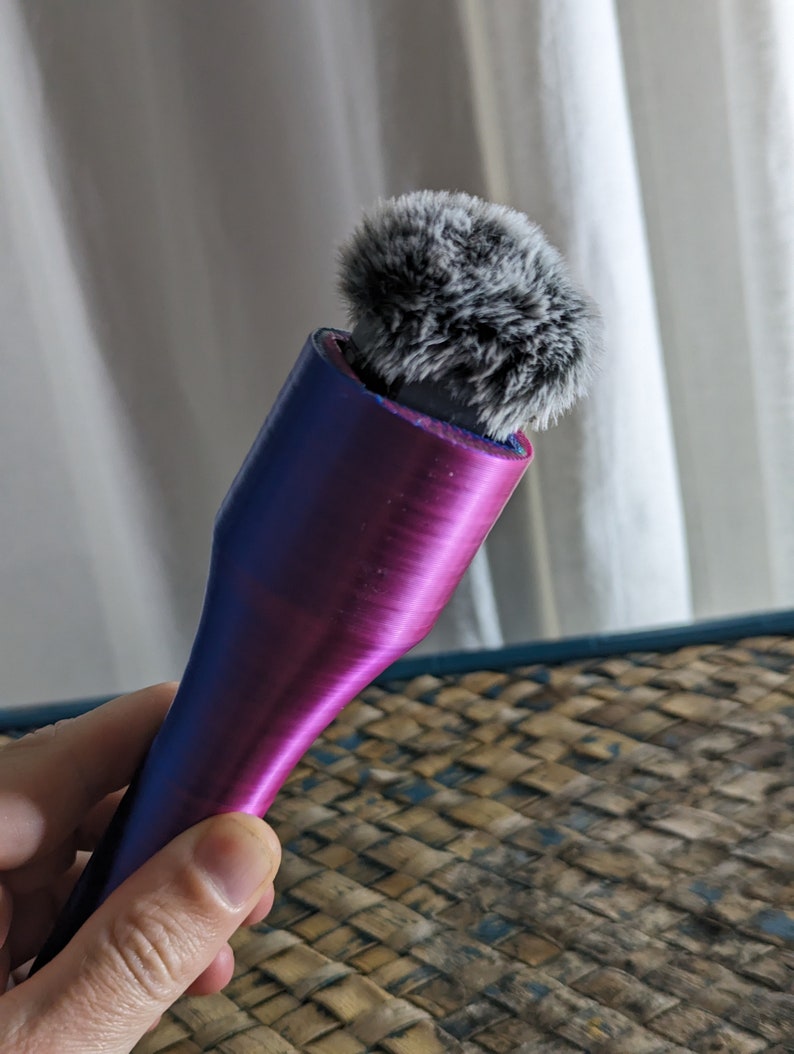 May include: A handheld microphone with a fluffy, gray windscreen. The microphone body transitions from blue to purple. The device is designed for recording audio. The background is a white curtain and a woven surface.