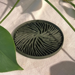 May include: A circular olive green soap dish featuring a raised, intricate, branching pattern that radiates from the center toward the outer rim. The design consists of numerous thin, curved ridges that create a textured, organic, and coral-like appearance.