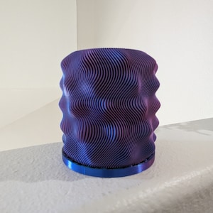 Op de afbeelding: Een cilindrisch decoratief object met een golvend, gelaagd ontwerp. De kleurovergang gaat van paars naar blauw, met een blauwe basis. Het object is gemaakt van meerdere dunne lagen, waardoor een textuur ontstaat. De algehele esthetiek is modern en artistiek.