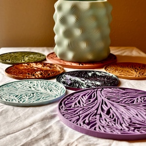 以下が含まれることがあります： ユニークで複雑なデザインのカラフルなコースター7枚セット。コースターは質感のある素材でできており、緑、茶色、紫、青など様々な色があります。コースターは、ライトブルーの波打つ円筒形のベースの周りに円形に配置されています。