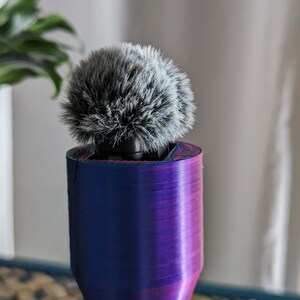 May include: A microphone with a fluffy, gray windscreen sits atop a cylindrical, gradient-colored stand. The stand transitions from blue to purple, with visible layered texture. The background includes a blurred plant and a neutral-toned wall.